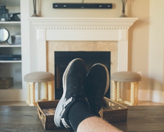A person is relaxing in a living room with feet up on a wooden table. A TV mounted above a fireplace is displaying a popular streaming platform. Shelves on either side of the fireplace hold decorative items. A smartphone can be seen on the table.
