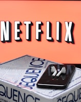 A screen displaying the Netflix logo in bold, black and white lettering against a vibrant red background. In front of the screen, there is a closed box for the board game 'Sequence', with a smartphone and wireless earbuds resting on top.