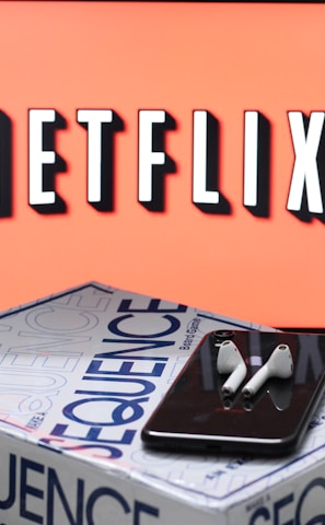A screen displaying the Netflix logo in bold, black and white lettering against a vibrant red background. In front of the screen, there is a closed box for the board game 'Sequence', with a smartphone and wireless earbuds resting on top.