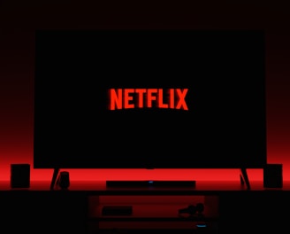 A television screen displays the Netflix logo in red against a black background. The room is dimly lit with a red glow possibly from LED backlighting behind the screen. Two speakers are positioned on either side of the TV set atop a minimalist entertainment center.