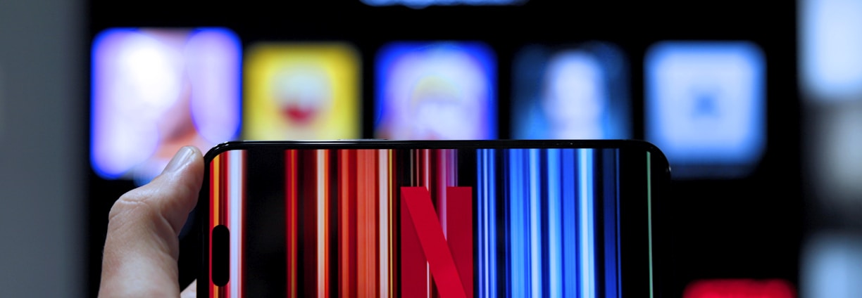 A hand holding a smartphone displaying the Netflix logo on its screen with a background of colorful vertical lines. In the background, a television screen shows blurry icons or profiles with various colors.
