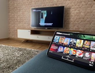 A modern living room features a flat-screen television mounted on a light-colored wooden stand with a patterned wall backdrop. Below the TV, a carpet lies on a wooden floor. In the foreground, a tablet displays a streaming platform interface with various show thumbnails, suggesting a relaxed entertainment setup.